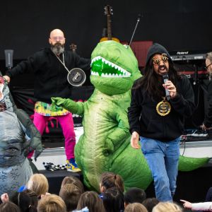 Mark Curtis rapping next to a green dinosaur and a grey dinosaur at a JAM Band show in Adelaide South Australia