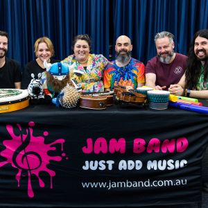 Elijah Maddern and Nina Verlingieri and Eve Ormsby and Nick Russell and Jono Ruse and Mark Curtis sitting behind a JAM Band banner with instruments in front of them in Adelaide South Australia