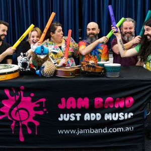 Elijah Maddern and Nina Verlingieri and Eve Ormsby and Nick Russell and Jono Ruse and Mark Curtis sitting behind a JAM Band banner and holding coloured boom whackers in Adelaide South Australia
